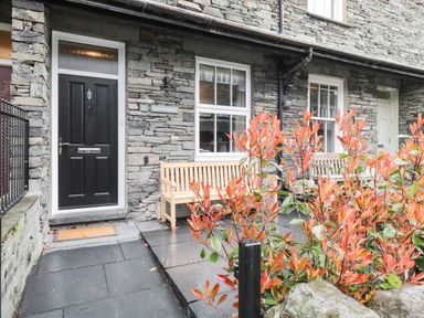 The front entrance of a stone house with a black door two wooden benches and a plant with orange leaves at Netherbeck in Ambleside