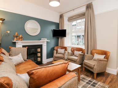 A living room with brown leather and fabric sofas a coffee table a fireplace with decorative stars and a round mirror a wall-mounted television and a window with beige curtains at Netherbeck in Ambleside