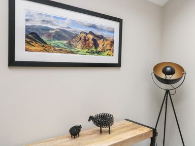A wooden table with two wire sheep figurines a framed landscape photograph on the wall and a tripod floor lamp in a corner at Netherbeck in Ambleside
