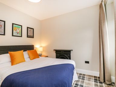 A bedroom with a bed with orange pillows a blue blanket a bedside table with a lamp framed pictures on the wall and plaid carpet at Netherbeck Ambleside