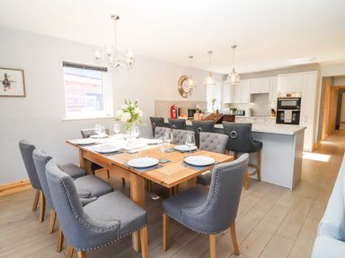 A dining area with a wooden table set with plates and glasses and grey upholstered chairs near a kitchen with bar stools at Lake View Lodge in Thorpe-On-The-Hill