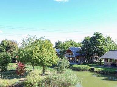 A pond with grass and trees surrounding wooden cabins at Lake View Lodge in Thorpe-On-The-Hill