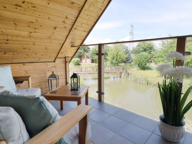 A cabin balcony with wooden furniture overlooking a pond and trees at Lake View Lodge in Thorpe-On-The-Hill