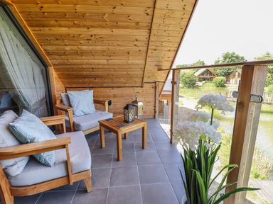 A balcony with wooden chairs cushions a small table lanterns and white flowers overlooking a pond at Lake View Lodge in Thorpe-On-The-Hill