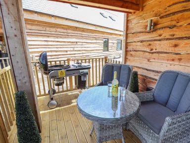 A covered wooden deck with a round glass table with a bottle and two glasses and two cushioned chairs at Lake View Lodge in Thorpe-On-The-Hill