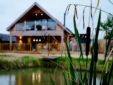 A wooden house with glass balcony behind tall grasses by a pond at Lake View Lodge in Thorpe-On-The-Hill