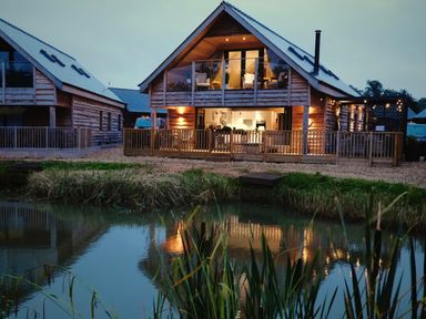 A wooden two-story house with a balcony and patio lights next to a pond at Lake View Lodge in Thorpe-On-The-Hill