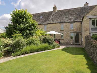 A stone house with a garden lawn and green bushes with patio furniture and an umbrella at Little Owl Barn in Burford