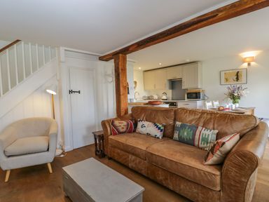 A living room with a brown leather sofa and patterned cushions a beige armchair a gray coffee table and a kitchen with dining table and flowers at Little Owl Barn in Burford