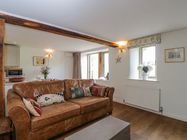 A living room with a brown leather sofa and colorful pillows near windows and a small kitchen at Little Owl Barn in Burford
