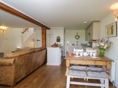 An open plan living area with a brown leather sofa dining table with chairs and bench kitchen with cabinets and a heart wall decoration at Little Owl Barn in Burford