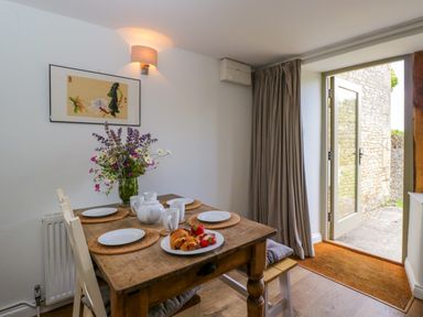 A dining area with a wooden table set with plates cups and pastries a glass door open to a stone patio at Little Owl Barn in Burford