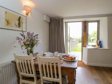 A dining area with a wooden table, white chairs, a vase of flowers, and a door leading outside at Little Owl Barn in Burford