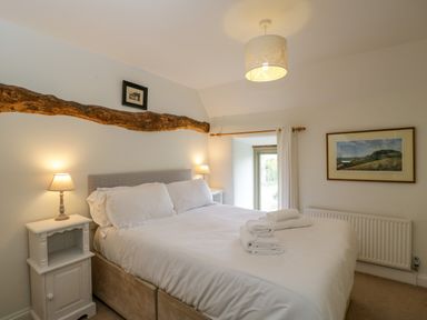 A bedroom with a large bed with white linens two bedside tables with lamps a wooden beam a window with white curtains and a landscape picture on the wall at Little Owl Barn in Burford