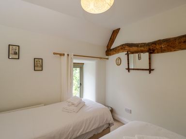 A bedroom with two beds with white linens a wooden beam a window with white curtains and framed pictures on the wall at Little Owl Barn in Burford