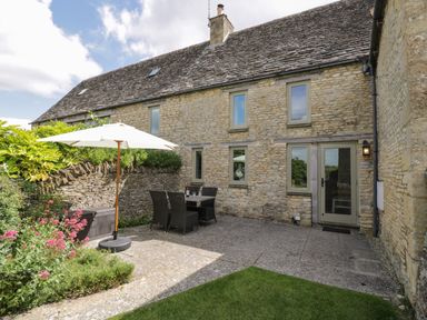 A stone patio with table chairs and a parasol outside a stone house with multiple windows at Little Owl Barn in Burford