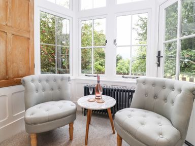 A corner with two upholstered chairs and a small round table with a bottle and two glasses by white windows at Eller Close House in Grasmere
