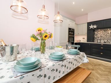 A dining table set with blue plates and glasses with flowers and a kitchen in the background at Eller Close House in Grasmere