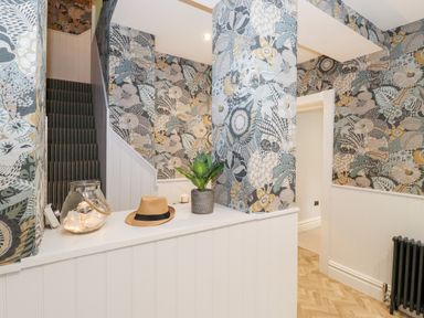 A hallway with patterned walls a staircase with striped carpet a plant in a pot a hat and a jar with lights on a white shelf at Eller Close House in Grasmere
