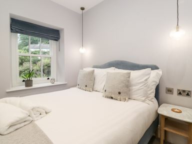 A bedroom with a double bed two pillows a bedside table with a coaster a window with a plant and an alarm clock at Eller Close House in Grasmere