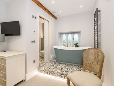 A bathroom with a freestanding bathtub on a tiled platform near a window a wooden chair a wall-mounted television and a small chest of drawers at Eller Close House in Grasmere