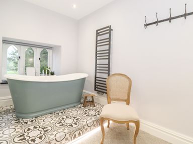 A bathroom with a green bathtub on patterned tiles a wicker chair and a black towel rack at Eller Close House in Grasmere