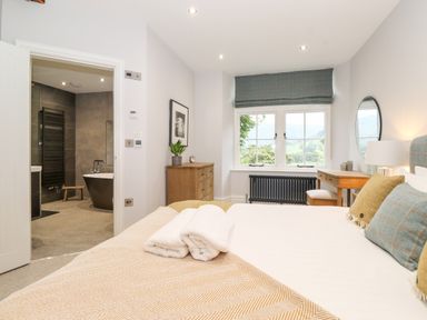 A bedroom with a bed, pillows, folded towels, a wooden chest of drawers, a desk with a stool, a round mirror, and a window overlooking greenery at Eller Close House in Grasmere