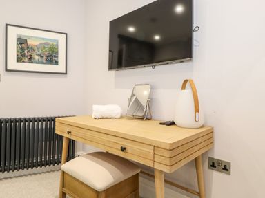 A wooden desk with a small mirror a lamp a rolled towel and a remote below a wall mounted television and a framed painting on the wall at Eller Close House in Grasmere