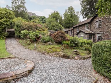 A stone cottage with a gravel driveway and garden with shrubs and trees at Eller Close House in Grasmere