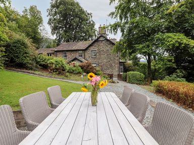 An outdoor wooden table with a vase of flowers and wicker chairs on a lawn in front of a stone house with trees and bushes at Eller Close House in Grasmere