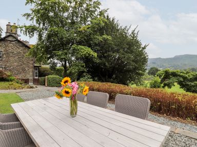 An outdoor wooden table with a vase of sunflowers and chairs in a garden with trees and a stone house in the background at Eller Close House in Grasmere