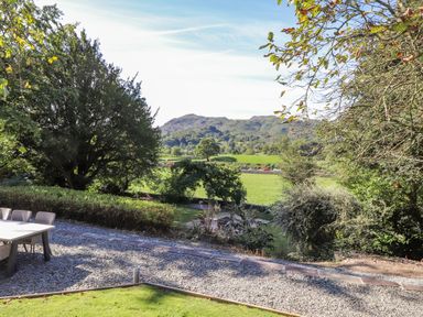 An outdoor seating area with a table and chairs on gravel near green trees and grass with hills in the background at Eller Close House in Grasmere