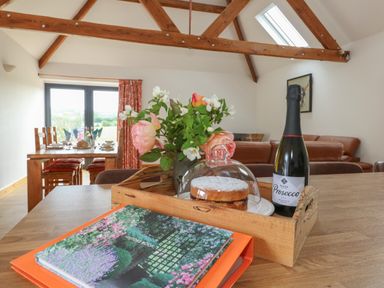 A wooden table with a tray holding a cake under glass, a bottle of prosecco, and a vase with flowers in a room with wooden beams and a dining area with chairs and a set table at Swallows Nest in Puxey near Sturminster Newton