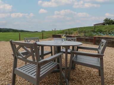 An outdoor seating area with a round table and four chairs on a gravel surface overlooking a grassy field at Swallows Nest in Puxey near Sturminster Newton