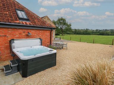 An outdoor hot tub next to a brick house with a table and chairs on gravel near a grassy field at Swallows Nest in Puxey near Sturminster Newton