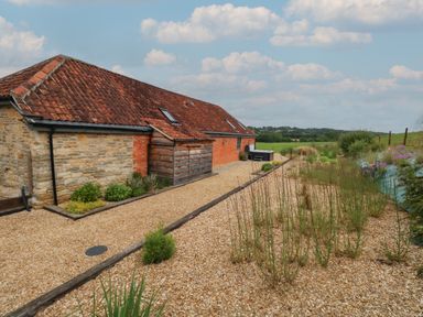 A stone and brick building with a tiled roof next to a gravel path and garden at Swallows Nest in Puxey near Sturminster Newton