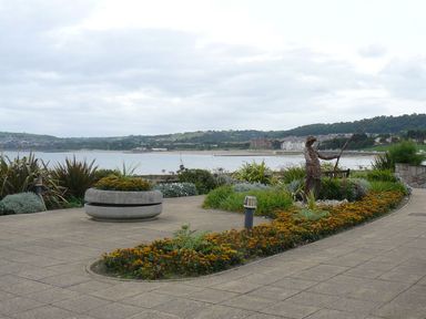 A waterfront promenade with flower beds and a fishing statue overlooking a bay with buildings in the distance at Ocean View Apartment in Colwyn Bay