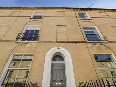The front facade of a three-story building with a grey door numbered 23 and windows with black railings at 23 Daniel Street in Bath