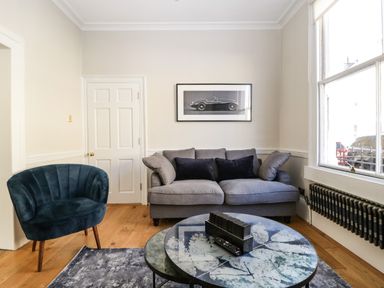 A living room with a gray sofa black pillows a blue chair round tables with a leaf design and a black and white car picture on the wall at 23 Daniel Street in Bath