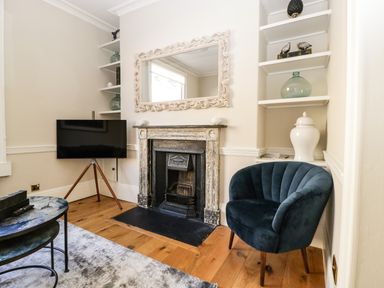 A room with a fireplace a mirror a tripod television a blue armchair and shelving with decorative objects at 23 Daniel Street in Bath