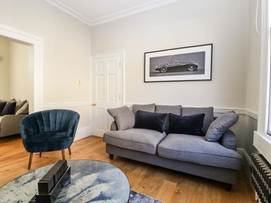 A living room with a grey sofa blue armchair round coffee table and a framed car picture on the wall at 23 Daniel Street in Bath