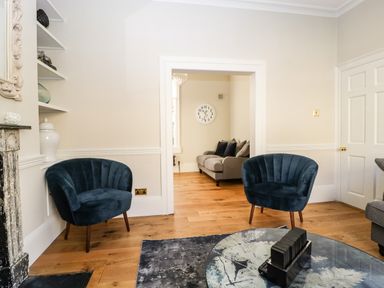 A living room with two blue chairs a grey sofa a clock on the wall wooden floor and a glass table at 23 Daniel Street Bath