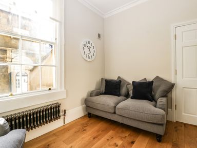 A corner of a room with a gray sofa with black cushions a white wall clock a large window and a radiator at 23 Daniel Street in Bath