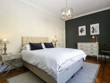 A bedroom with a large bed, bedside tables with lamps, a dresser with a lamp, two framed pictures on a dark wall, and a chair at 23 Daniel Street in Bath
