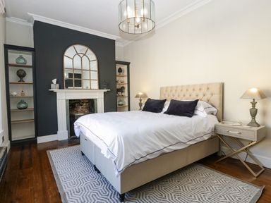 A bedroom with a beige bed and white bedding dark pillows beside a fireplace with a mirror and shelves at 23 Daniel Street in Bath