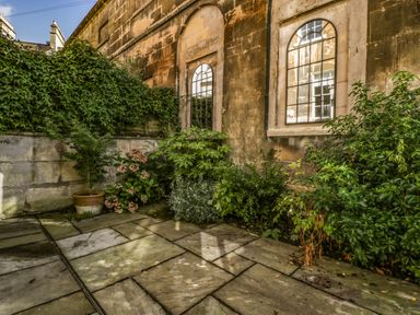 An outdoor paved area with plants and shrubs along walls with arched windows at 23 Daniel Street in Bath