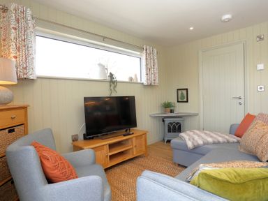 A living room with a gray armchair and sofa a TV on a wooden stand and a window with patterned curtains at The Wren in Wem