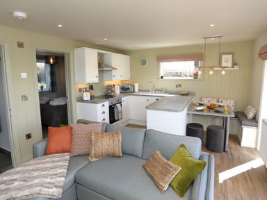 An open living space with a grey sofa and cushions next to a white kitchen with a dining nook at The Wren in Wem