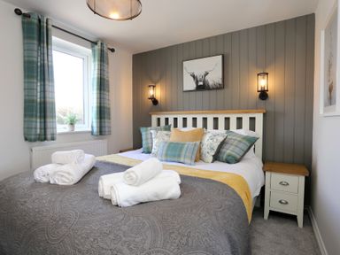 A bedroom with a bed with pillows and folded towels by a window with plaid curtains at The Wren in Wem