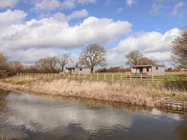 Two wooden cabins with porches behind a fence by a river with trees in the background at The Wren in Wem